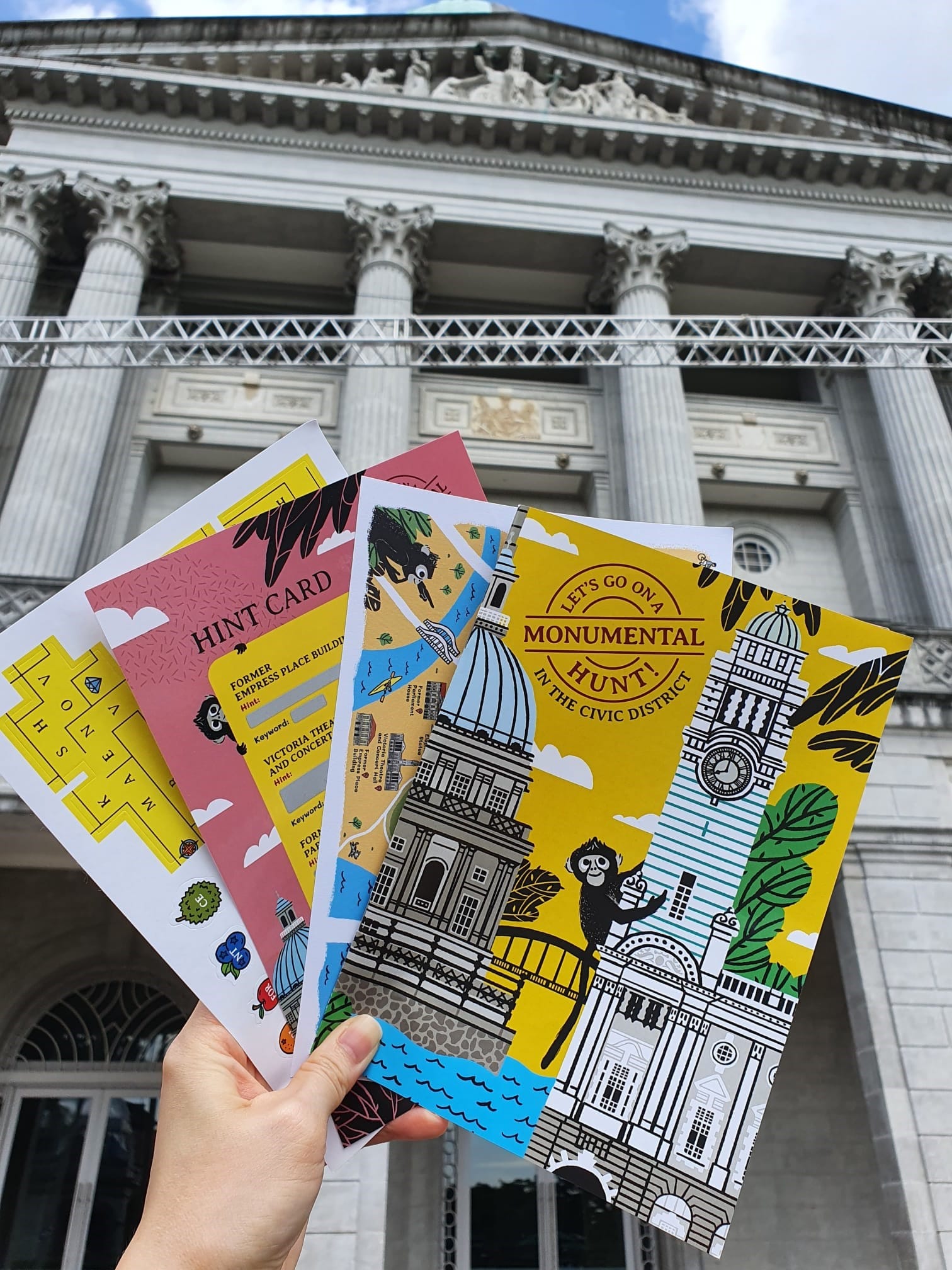 Hand holding colorful activity cards for a Monumental Hunt in the Civic District in front of a grand columned building.